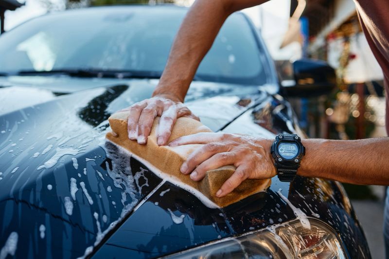 Close-up of hands washing a black car using a large sponge covered in white soap suds. Outdoor setting with sunlight reflecting on the vehicle’s glossy surface during daytime.
