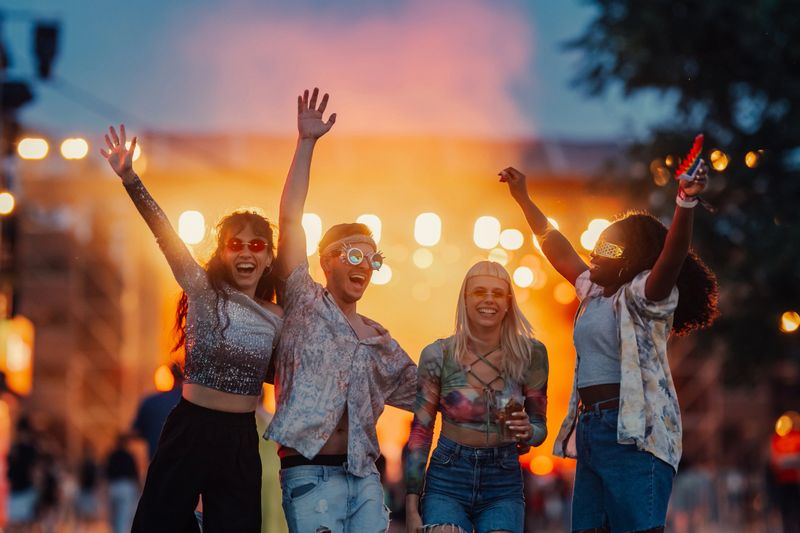 Four friends are raising their arms and dancing at a music festival, enjoying the vibrant atmosphere and live performance