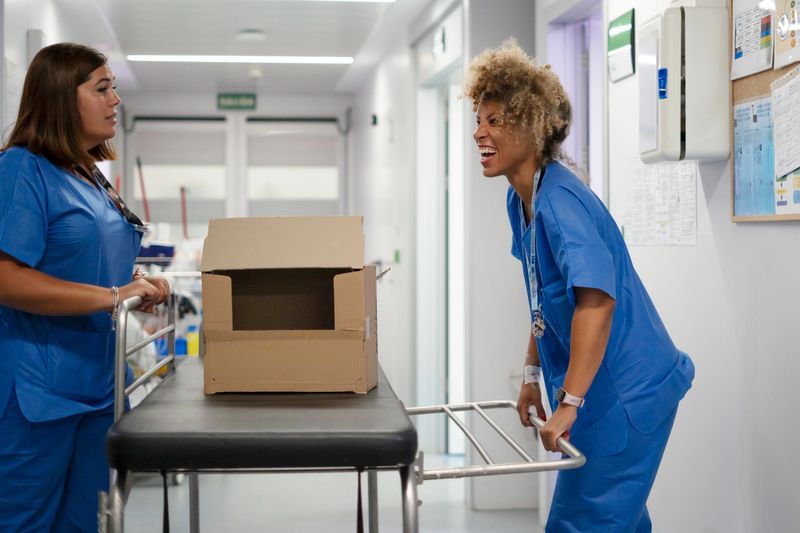 Two nurses transporting a cardboard box on a gurney through a hospital corridor, laughing at work
