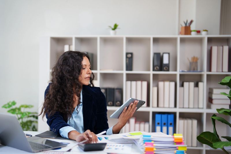 Professional businesswoman calculating finances and managing digital information using a digital tablet at a busy office desk, demonstrating multitasking and productivity
