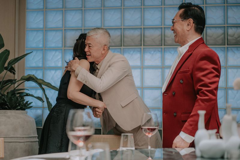 chinese senior man warmly hugs an Asian woman during a reunion at an upscale restaurant while an Asian middle-aged man smiles nearby, expressing friendship, celebration, and luxury dining lifestyle. Kuala Lumpur
