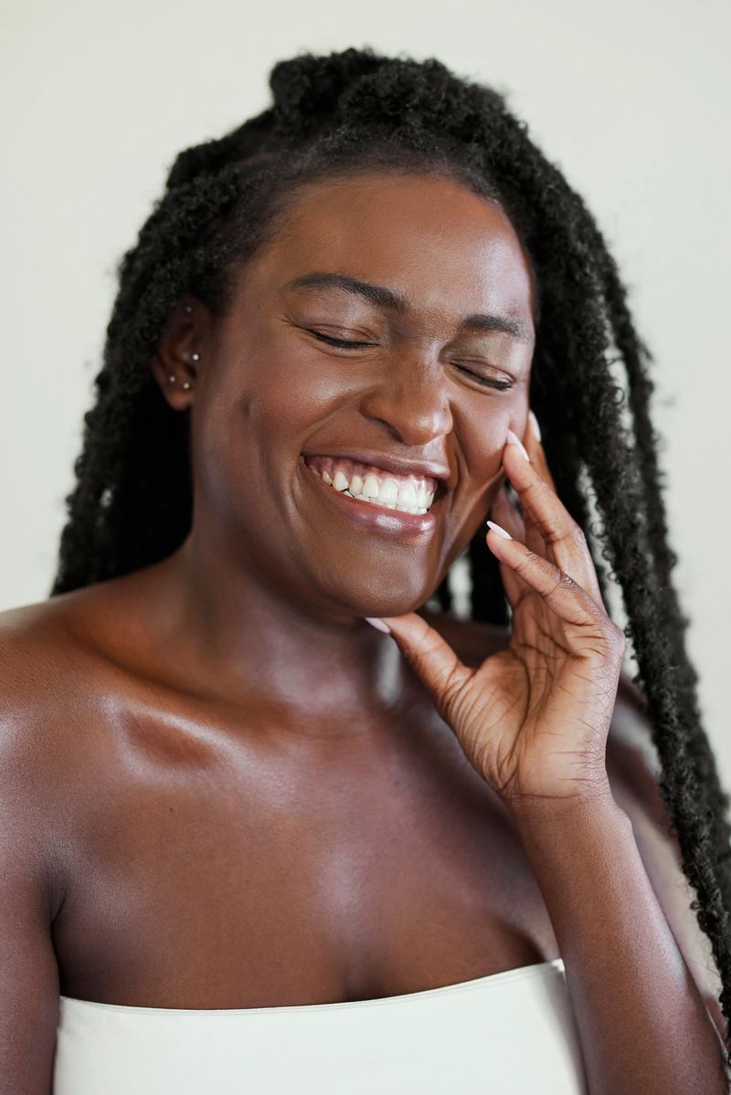 Vertical beauty portrait of a joyful woman with dark skin and long hair twists laughing with her eyes closed while touching her glowing face.