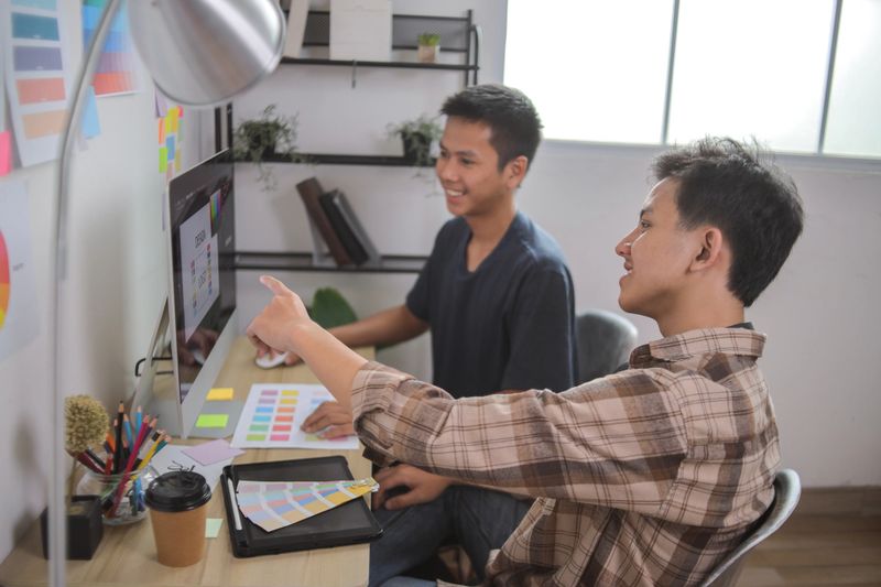 Two Smiling Graphic Designers Collaborating in Office, Pointing at Computer Screen While Selecting Colors From Physical Swatch