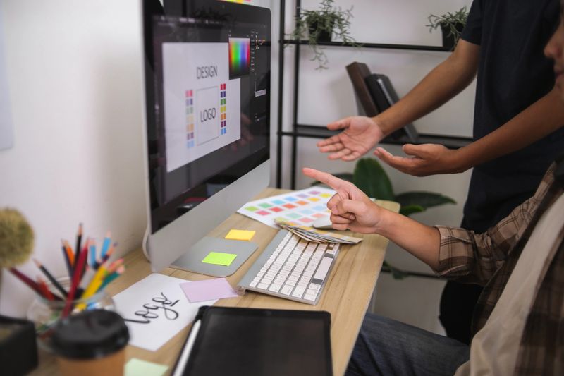 Two Graphic Designers Collaborating on Monitor, Reviewing Color Palettes and Logo Design Concepts in Modern Office