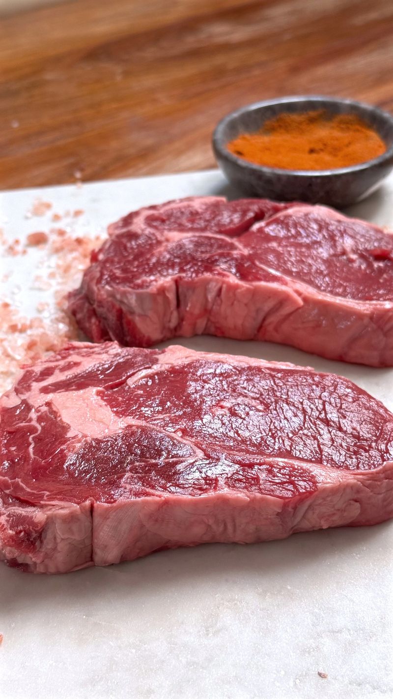 Two Strip Steaks With Smoked Paprika And Herb Sprigs On Marble, Chef Tasting Prep For Tasting Menu, Refined Seasoning And Precise Portioning, Elevated Restaurant Ambiance