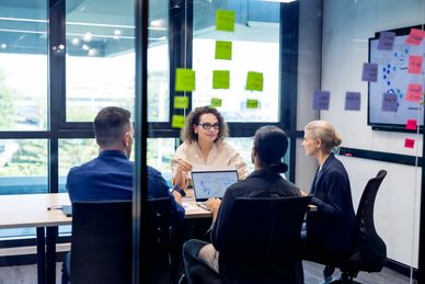 Four people in a meeting room with sticky notes on the glass wall.
