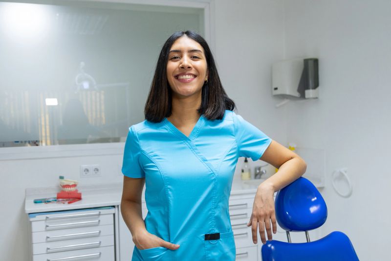 Young woman in medical scrubs smiling at camera, leaning on a dental chair in a modern clinic setting