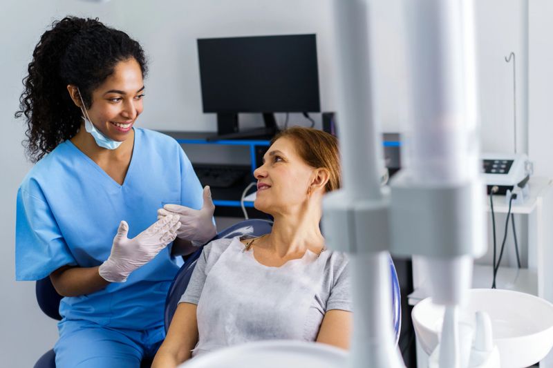 A smiling nurse in blue scrubs and gloves converses with a seated patient in a dental clinic, conveying care, comfort, and professional healthcare support.