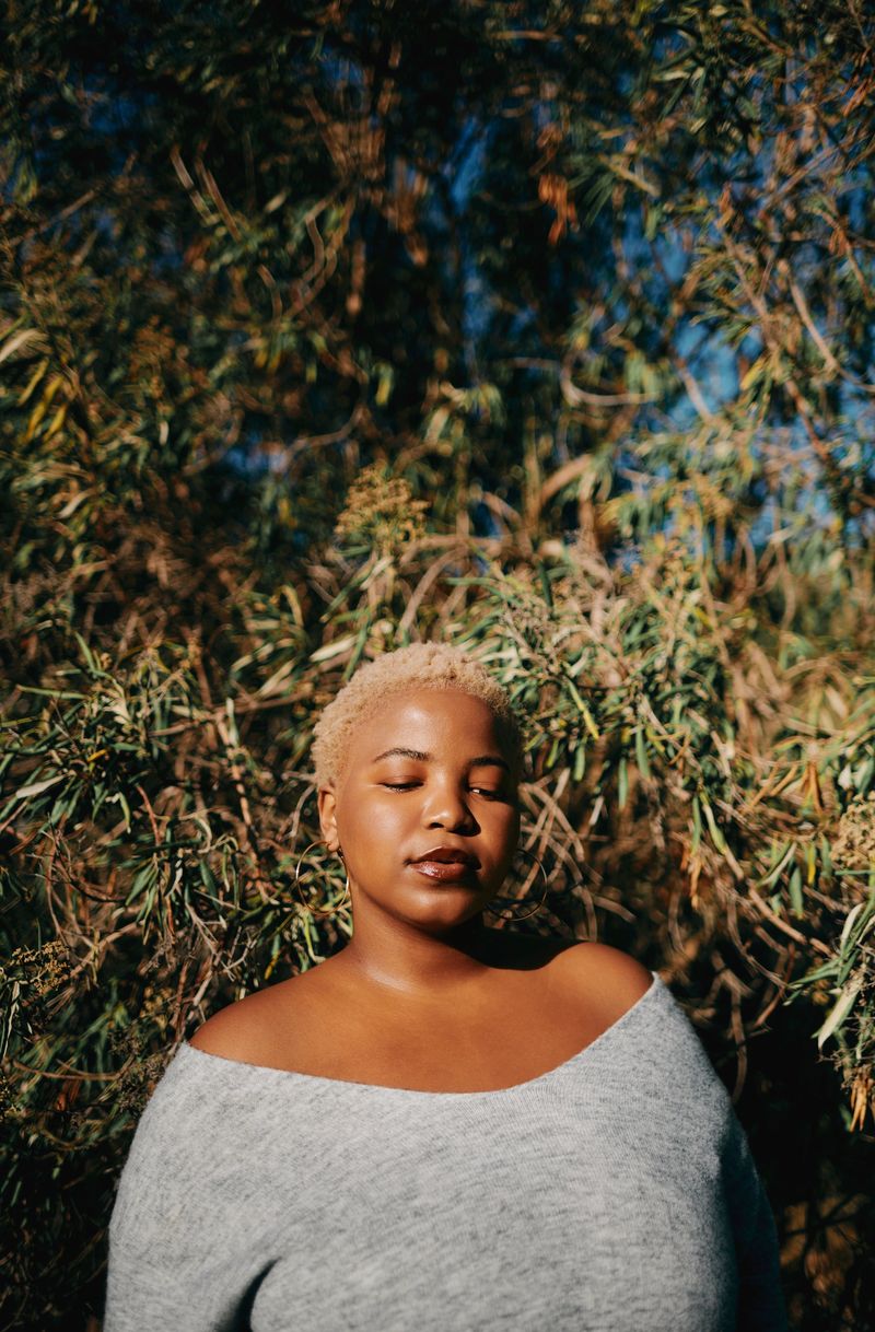 A young Black woman with short blonde hair wearing a gray off-shoulder sweater stands outdoors surrounded by green foliage, eyes closed, embodying calm and digital detox in natural sunlight