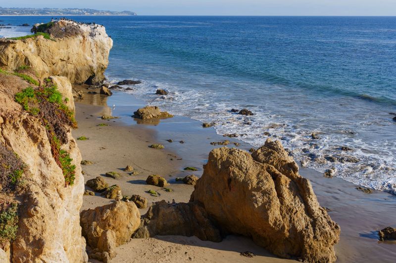 Rugged sea stacks and expansive sandy beach define El Matador State Beach in Malibu, with tranquil ocean waves gently meeting the shore under a bright sky.
