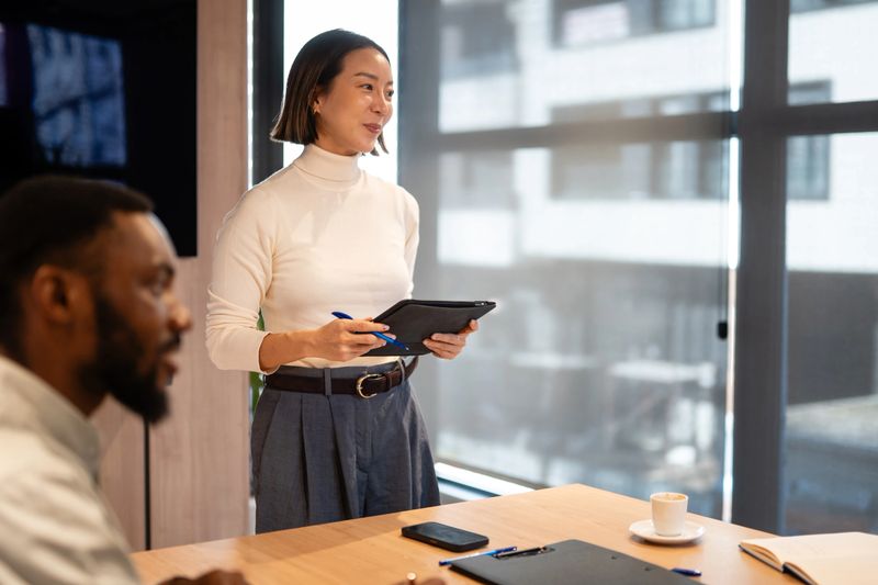 Asian business woman standing, holding a tablet and a pen, leading a presentation during a corporate team meeting in an office