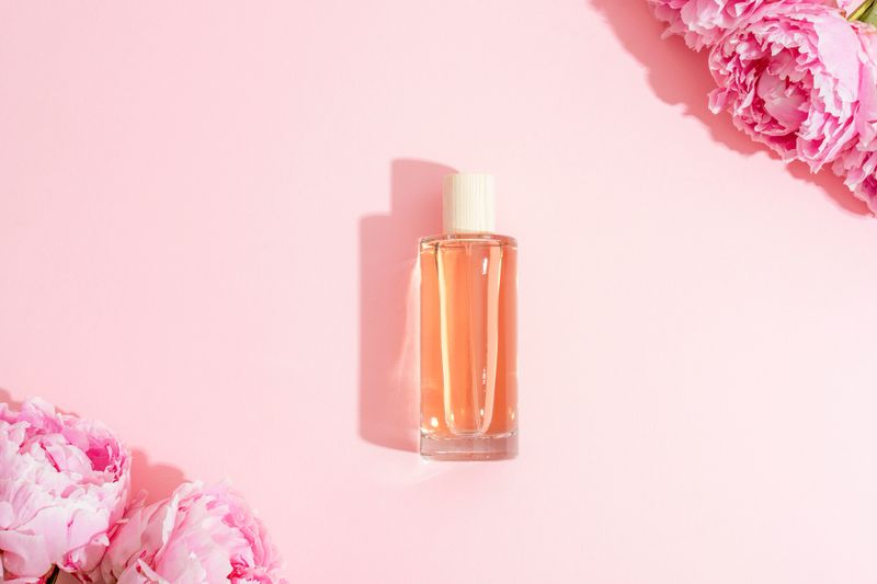 Glass perfume bottle with soft pink liquid surrounded by peony flowers on pastel pink background. Top view, flat lay.