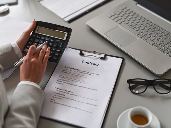 Person calculating contract details at a desk with laptop, glasses, and coffee.