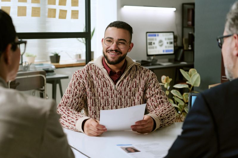 Young adult man smiling while holding resume, participating in job interview with two middle aged professionals in modern office setting, engaging in conversation