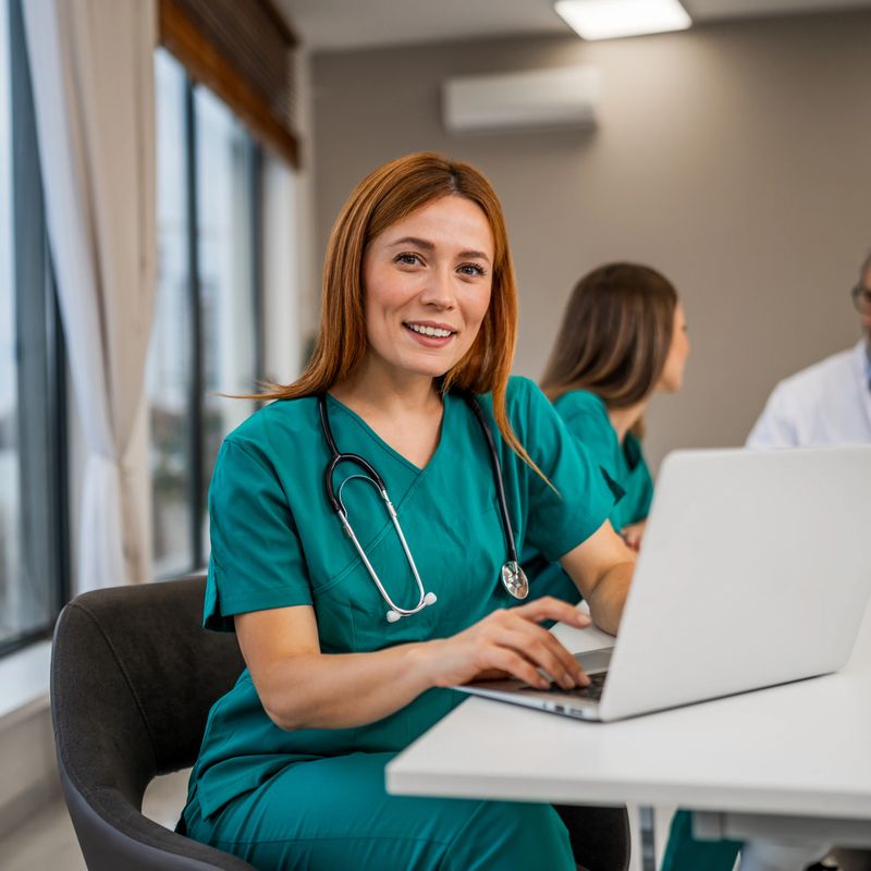 Nurse smiling while working on a laptop at a table with colleagues in the background, representing healthcare collaboration