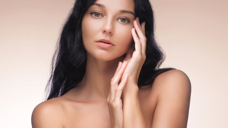 A stunning woman with long dark hair touches her face with a serene expression, emphasizing her flawless skin against a beige background. Perfect for beauty, skincare, and self-care themes.