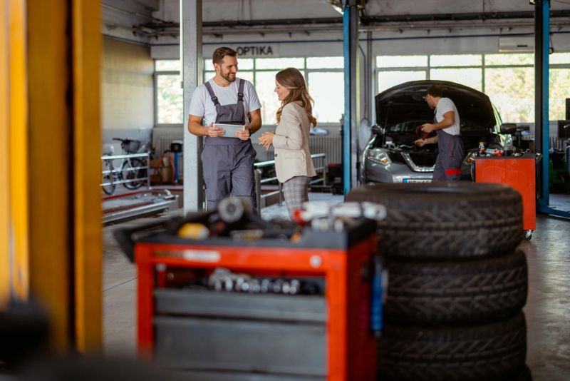 A mechanic consults with a client against the backdrop of a fully equipped garage, where a second technician is diligently working on a car. The scene conveys trust, skill, and the customer-centric approach of a modern repair shop.