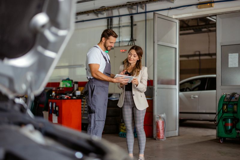 A mechanic in a modern repair shop uses a digital tablet to consult with a colleague or client. The workspace is surrounded by tools and equipment, emphasizing innovation, precision, and top-notch customer care. The composition effectively conveys themes of customer service, collaboration, and technical expertise in the automotive maintenance industry.