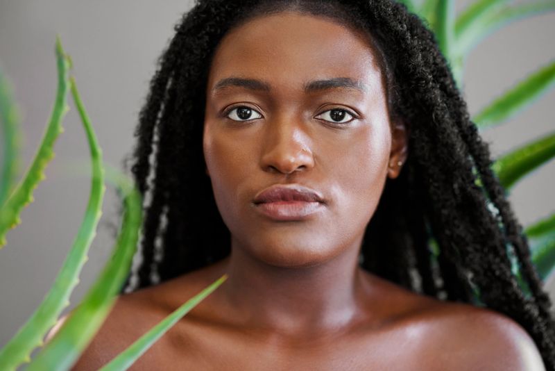 A serene portrait of a young Black woman with radiant skin and long braids, framed by green aloe vera leaves in a beauty and wellness concept.