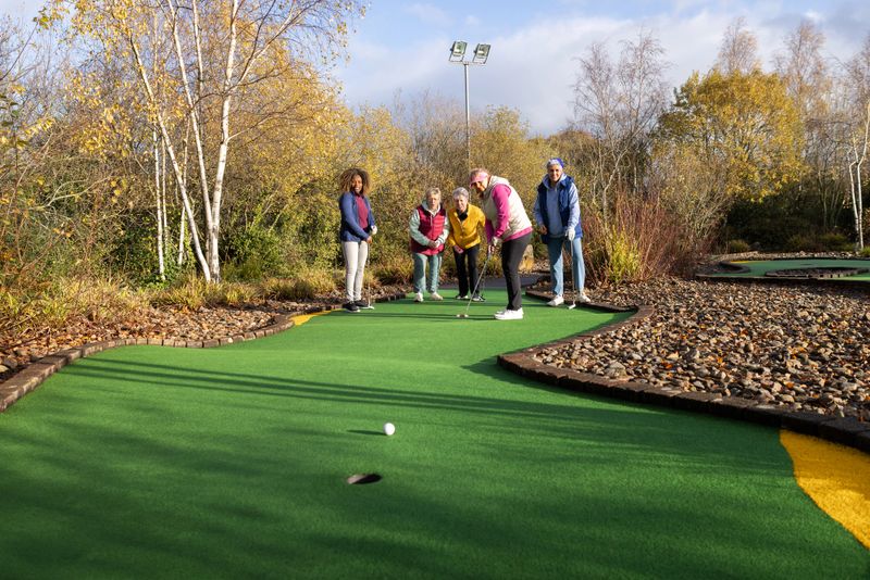 Wide shot of a small group of mature females playing a game of mini golf at a golf club in Newcastle-upon-Tyne, North East England. One person is taking a shot while the others all watch in anticipation as the ball rolls towards the hole. It is a sunny winter's day and they are wearing warm sportswear.Videos similar to this scenario available.