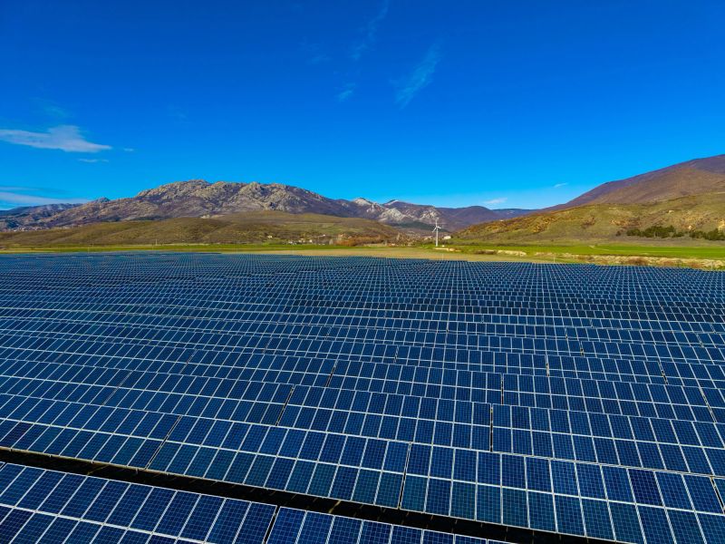 Rows of solar panels stretch across the land, collecting sunlight for energy. Mountains stand in the background under a clear blue sky, showing a focus on renewable energy.