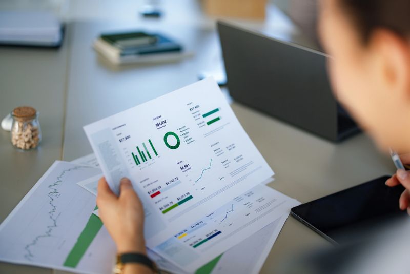 A professional analyzes printed financial charts and graphs at a bright office desk, surrounded by a laptop, tablet, and documents. The scene conveys data-driven decision making and corporate planning.