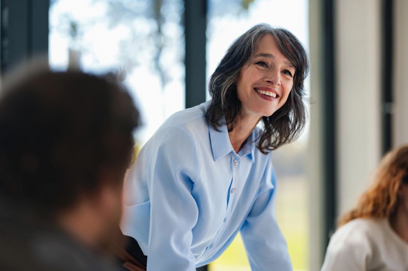 A cheerful woman in a light blue shirt leans over a desk, engaging with colleagues in a sunny office. The mood is positive, collaborative, and professional, conveying teamwork and open communication.