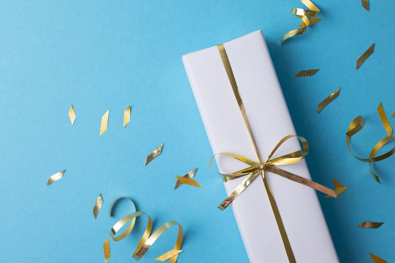 White Gift Boxes With Golden Ribbons, Top View On Beige Background Scattered With Gold Confetti And Curled