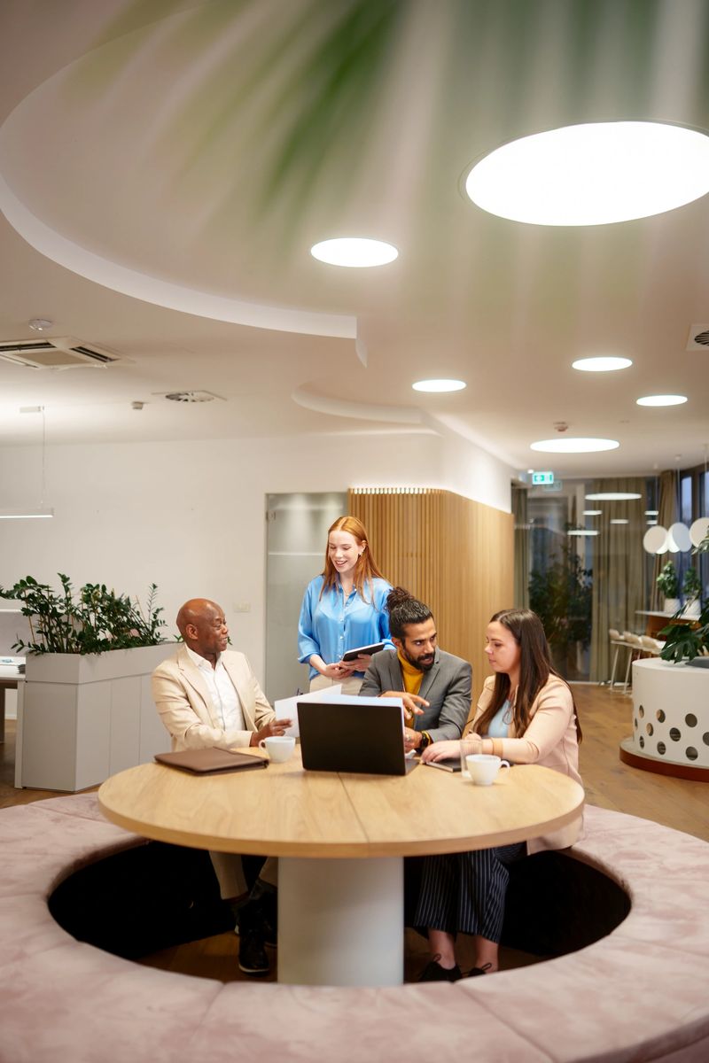 Diverse young and mid adult businesspeople brainstorming digital content in a cozy office, collaborating around a modern round table. They are actively discussing project details, utilizing a laptop and a digital tablet for data review and planning.