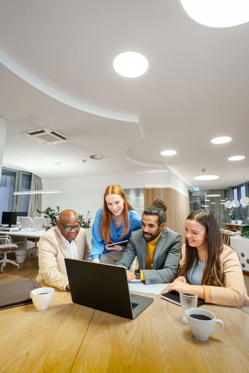 Business professionals in a cozy office brainstorming around a laptop. Group is engaged with digital technology, collaborating on a project. Interior features modern furniture and lighting. Daytime setting.
