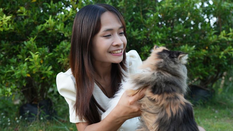 Woman smiles while holding fluffy cat outdoors, surrounded by greenery. joyful interaction highlights bond between them