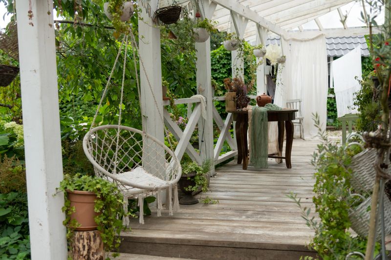 A wooden deck with a swing chair and a table is present in a garden area. Green plants hang from the ceiling. The scene shows a bright daylight setting with various plants around.