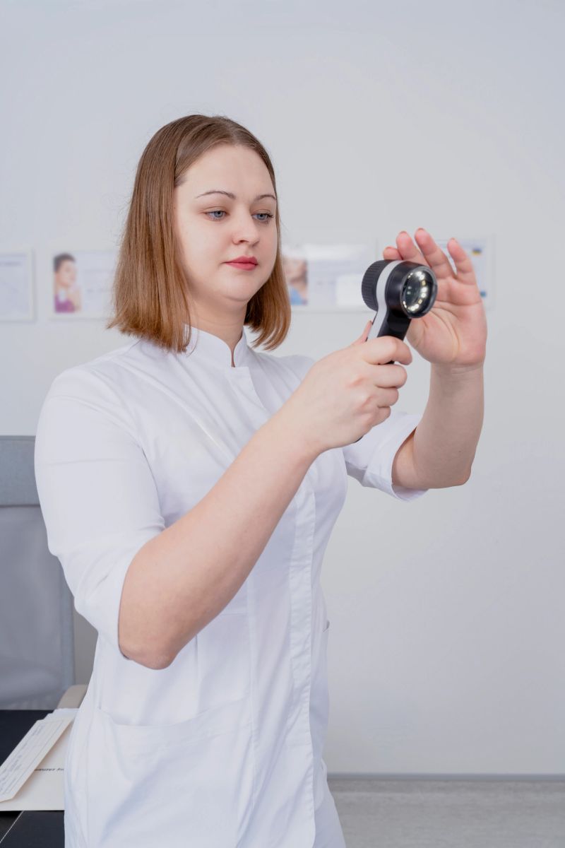 Female healthcare worker inspects skin using a dermatoscope in a clinical environment, with medical charts visible on the wall behind her