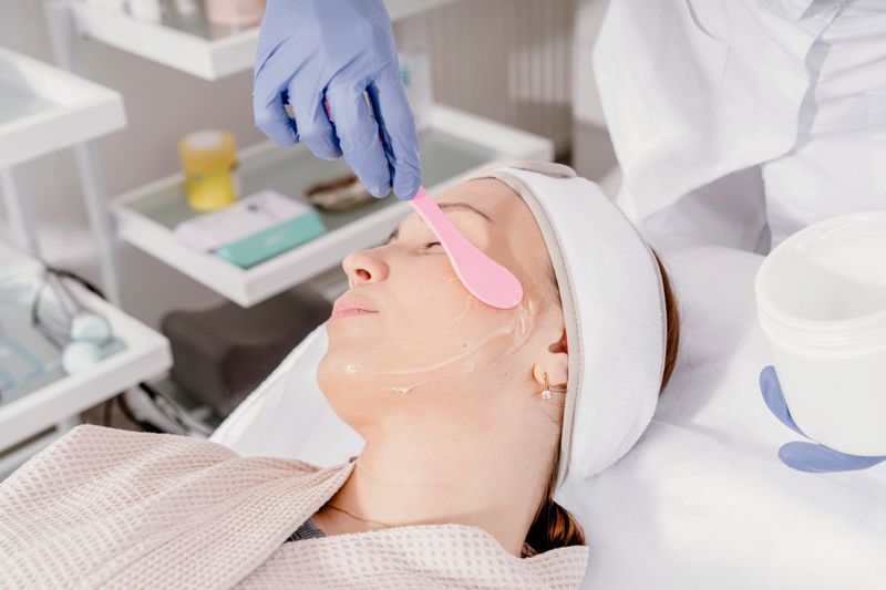 Woman undergoing a facial treatment in a spa, esthetician using a pink spatula to apply cream on her face while she lies on a treatment table
