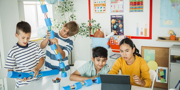 Children building a model rocket while studying on a tablet in a classroom.