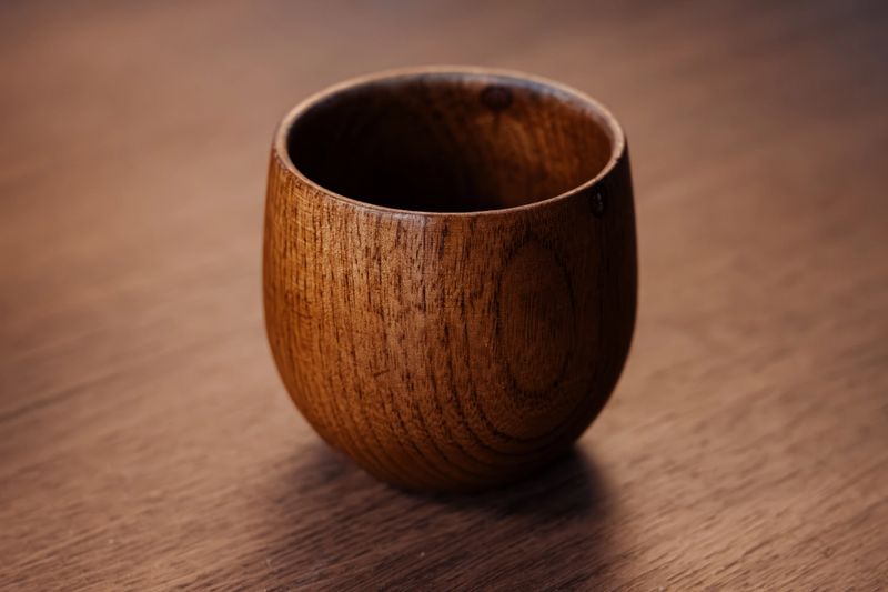 A detailed shot of a single, handmade wooden cup placed on a rustic wood surface, emphasizing its natural grain and warm texture. Ideal for themes of sustainability, craftsmanship, and home decor.