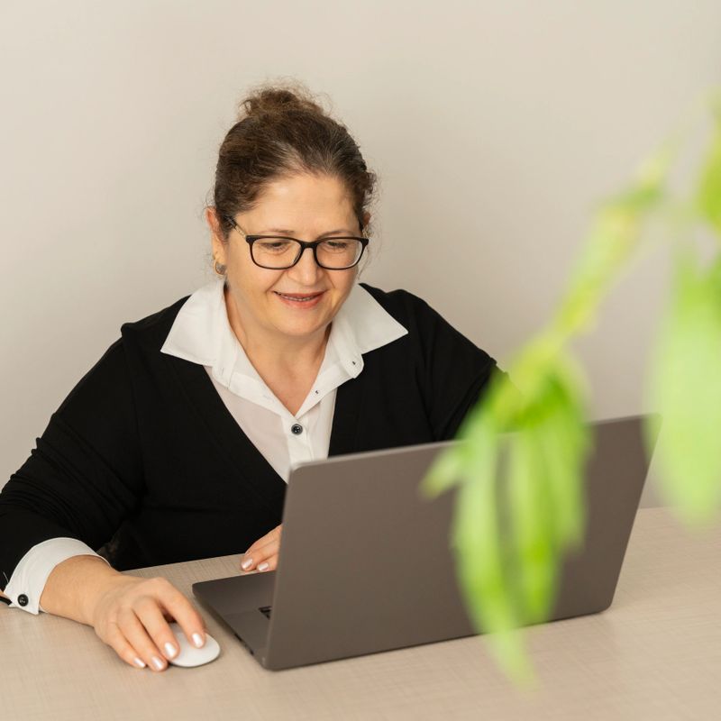 Portrait of a cheerful mature woman sitting at a desk, looking at her laptop screen with a bright smile.