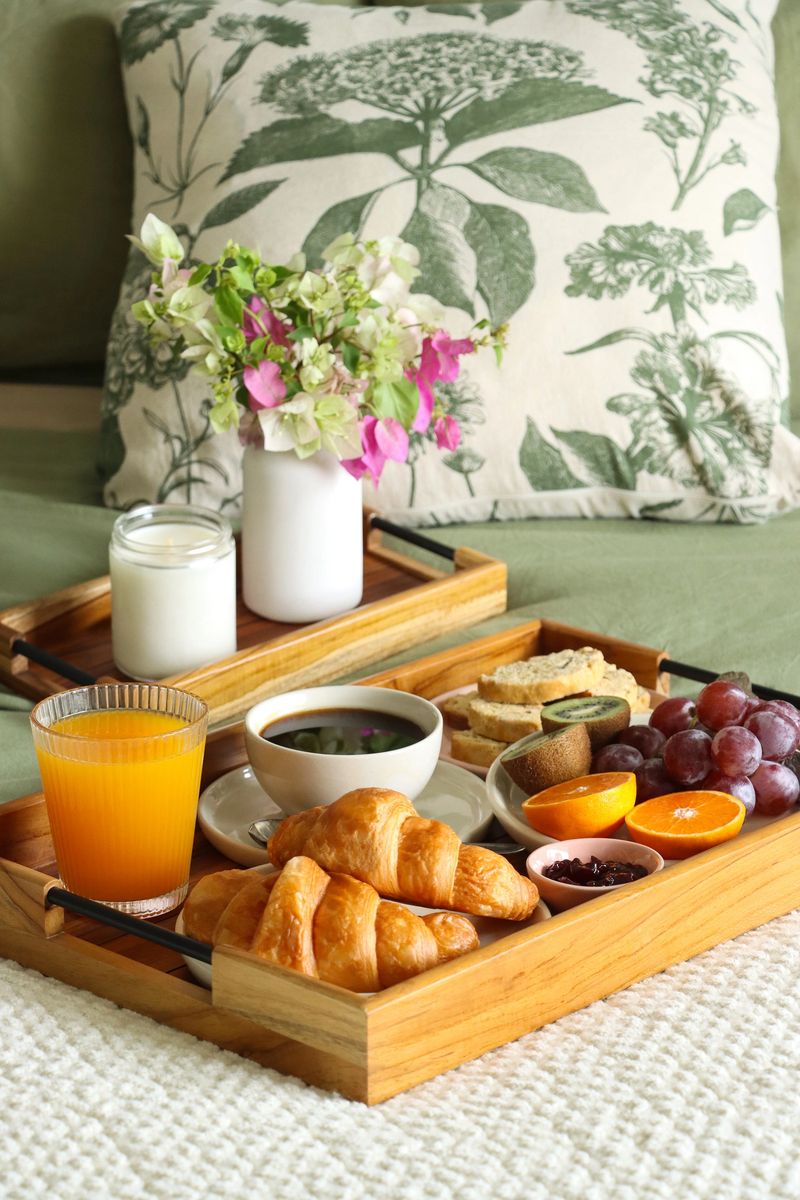 Stock photo shows a romantic breakfast in bed arranged on a wooden tray featuring orange juice, croissants, pastries, biscotti, fresh fruit, flowers, and coffee. This cozy and inviting setup highlights indulgent morning rituals, comfort, and elegance