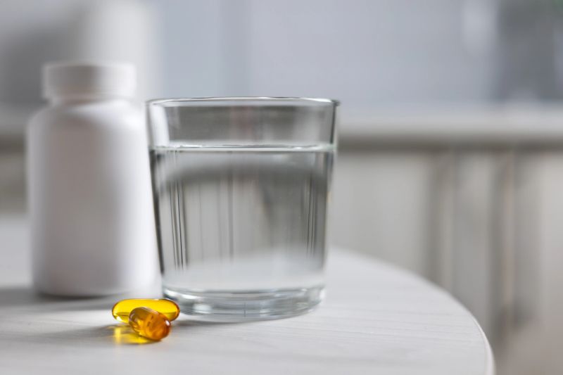 Omega Capsules Beside White Bottle On Round Table With Glass Of Water, Softgel Sheen Reflecting Morning Light,