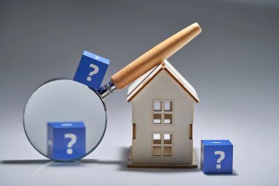 Magnifying glass, wooden house model, and blue question mark cubes symbolizing real estate uncertainty.