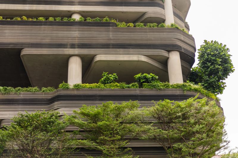 Rows of plants decorate the balconies of a tall building. The structure has a unique design with lines and pillars. Trees are in front of the building, adding greenery to the scene.
