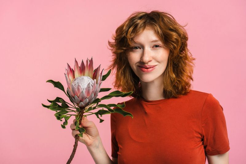 Portrait of smiling beautiful young woman holding flower protea looking at camera isolated on pink background. Advertisement concept