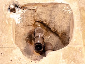 Exposed plumbing pipes in a floor hole with damaged tiles.