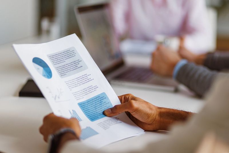 A group of professionals in a business meeting engage in a brainstorming session, examining financial documents and using a laptop to explore ideas. The image captures a modern office environment and teamwork.
