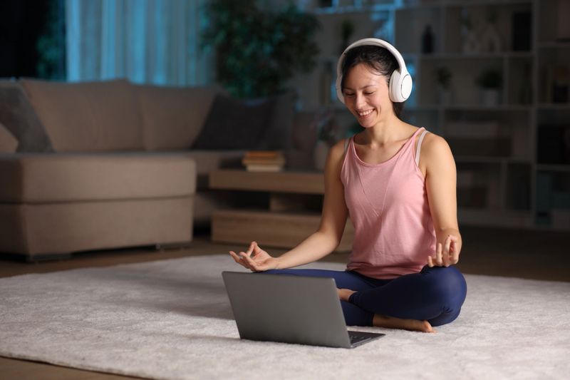 A Brown woman smiling warmly while following an online wellness class on her laptop from a cozy, plant-filled home.
