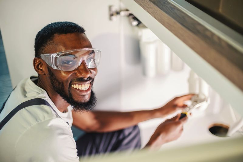 Plumber wearing safety goggles and overalls, using a wrench to repair pipe fittings under a kitchen sink while smiling at the camera, providing professional home maintenance service