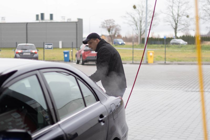 A man uses a pressure washer wand to clean the rear bumper of a car at a car wash.