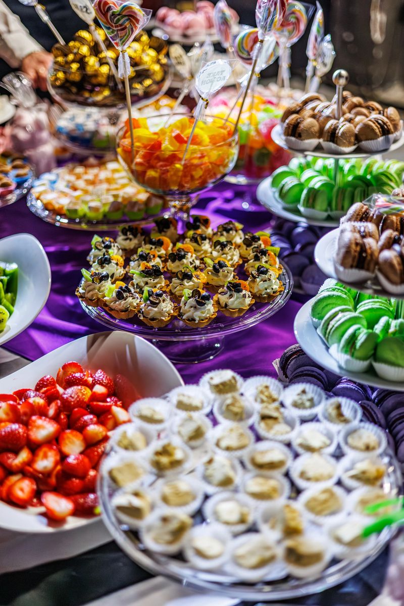 A vibrant dessert table featuring green macarons, mini tarts, lollipops, strawberries, and assorted sweets on elegant trays over a purple tablecloth