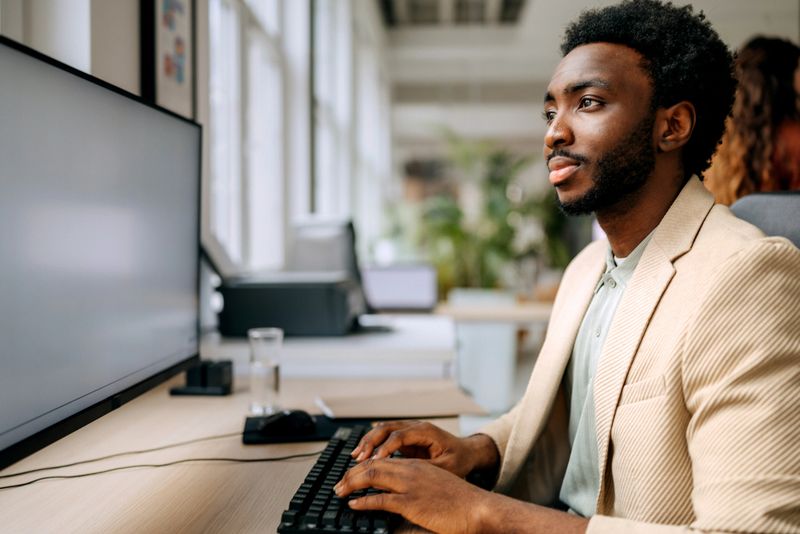 In a modern office setting, a young person sits at a desk working on a computer with a focused expression. Others are present in the background, engaged in tasks.