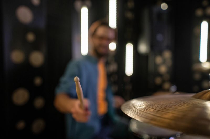 Musician playing drums with a drumstick on a cymbal, creating rhythm and sound during a performance in a recording studio with blurred lights in the background